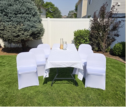 Six-foot table dressed in white linen with fitted white chair covers on a lawn